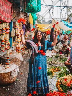 a woman standing in front of a market with lots of flowers on the ground and decorations hanging above her