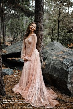 a woman standing next to a tree in the woods wearing a long pink dress and smiling