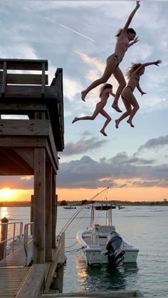 two women jumping into the water from a dock at sunset with boats in the background