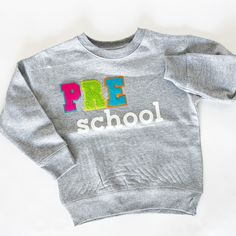 Our pre-k mini chenille patch sweatshirt is perfect for any little one with school spirit.  Letters are available in both a white-black option and a multi-color option for a fun youthful look.   Sweatshirt runs true to size for children. It's suggested women size down for a more fitted look. Women keep your traditional size for a relaxed/oversized fit.  8-ounce, 50/50 cotton/poly Double-needle stitching at waistband and cuffs 1x1 rib-knit collar, cuffs, and waistband with spandex Sporty Letter Embroidery Sweatshirt For School, School Spirit Tops With Letter Embroidery, Varsity Cotton Tops For School, Cotton Varsity Tops For School, Cotton Crew Neck Tops For School Events, Crew Neck Cotton Tops For School Events, Long Sleeve Tops For End Of School Year Events, Long Sleeve Tops For School Events, Long Sleeve Sweatshirt With Embroidered Logo For School