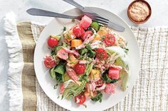 a white plate topped with a salad next to a fork and bowl filled with dressing