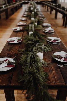 a long wooden table with plates and silverware on it, along with greenery