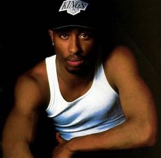 a man wearing a white tank top and a black hat sitting on a chair with his arms crossed