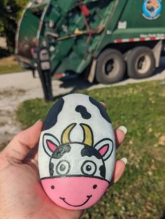 a hand holding a painted rock with a cow on it and a garbage truck in the background