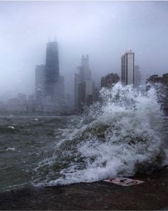 an ocean wave crashes into the shore in front of a large city on a foggy day