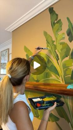 a woman is painting a wall with green leaves on it and she is holding a paintbrush in her hand