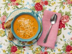 a bowl of soup with cheese on top next to a spoon and napkin, sitting on a floral tablecloth