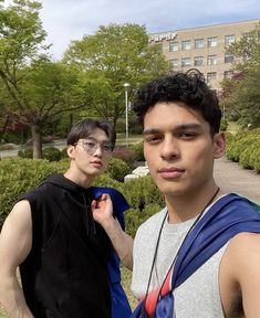 two young men standing next to each other in front of trees and bushes with buildings behind them