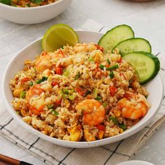 a white bowl filled with rice and shrimp next to cucumbers on a table