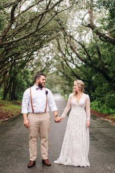 a bride and groom holding hands while walking down the road in front of some trees