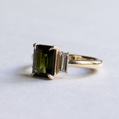 Emerald cut green tourmaline adorned with baguette diamonds Metal: 14K yellow gold Stone: Green Tourmaline Stone Size: 8 x6 mm Stone Weight: 1.7 Carat Accent Stone: Baguette Diamonds: Diamond Size: 4.5 x 2 mm (2) Diamond Color and Clarity: GHI, SIDiamond weight: 0.12 carat each Total Diamond Weight: 0.25 carat Gold Engagement Ring Three Stone, Engagement Ring Three Stone, Emerald Cut Ring, Smaragd Ring, Ring Three Stone, Trio Ring, Emerald Cut Rings, Baguette Diamonds, Tourmaline Stone