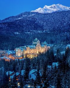 a large building surrounded by trees and snow covered mountains in the distance is lit up at night