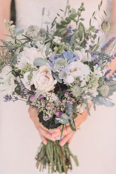 a woman holding a bouquet of flowers and greenery in her hands while wearing a white dress