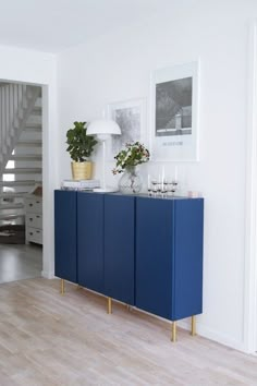 a blue cabinet with gold legs in a white room next to a lamp and pictures on the wall