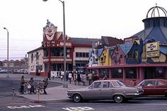 cars parked on the street in front of a building with people walking around and riding bikes