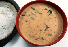 a red bowl filled with soup next to a piece of bread on a white surface