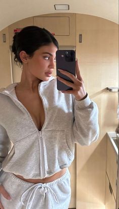 a woman taking a selfie with her cell phone in a bathroom mirror wearing a gray hoodie and matching sweatpants