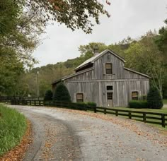 an old barn sits on the side of a gravel road in front of a wooden fence