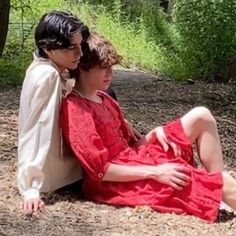 two people are sitting on the ground in the woods, one is wearing a red dress