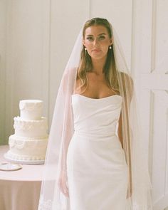 a woman standing next to a white cake on top of a table with a veil over her head