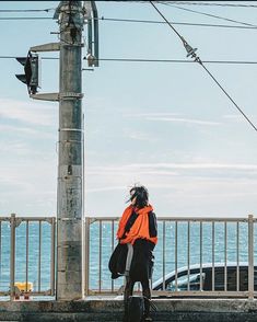 a woman is walking down the street by the ocean with an orange jacket and black pants