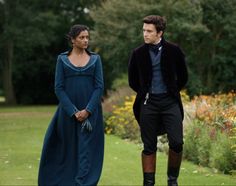 a man and woman dressed in period clothing walking through a garden with flowers behind them