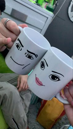 two coffee mugs with faces drawn on them being held by someone's hand