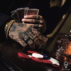 a tattooed hand holding playing cards on top of a table next to a bottle and lighter