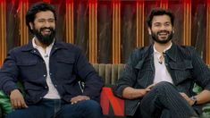 two men sitting on couches in front of a red and yellow curtain, one is smiling at the camera