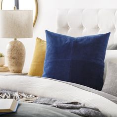 a bed with blue and yellow pillows on top of it next to a book, lamp and mirror