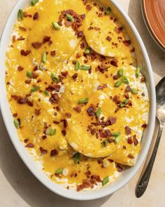 a white bowl filled with cheese and bacon covered in green onions next to silverware