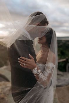 a bride and groom embracing each other under a veil on top of a cliff overlooking the ocean