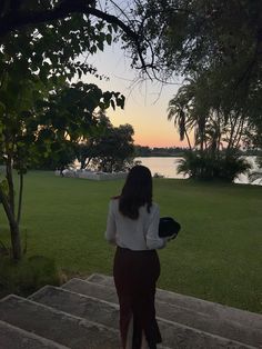 a woman is walking up some steps towards the water at sunset or dawn with her hat in hand