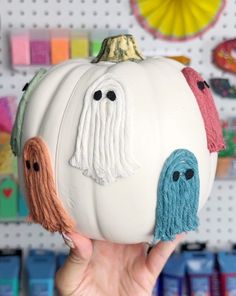 a hand holding up a white pumpkin with two faces painted on it and other decorations in the background