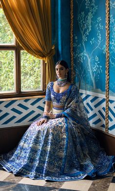 a woman sitting in front of a window wearing a blue and white dress with gold trim