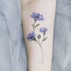 a small blue flower tattoo on the left forearm and foot, with green leaves growing out of it