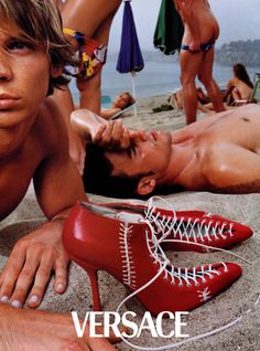 a man laying on the beach next to a pair of red high heeled shoes