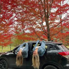 two women are upside down on the hood of a car in front of trees with red leaves