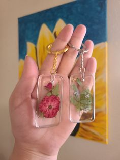 a person holding two keychains with flowers on them in front of a painting