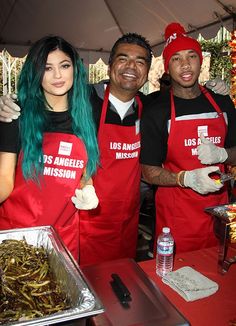 three people wearing red aprons and green hair stand in front of a table with food