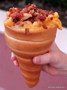 a person holding an ice cream cone filled with macaroni and cheese covered in bacon