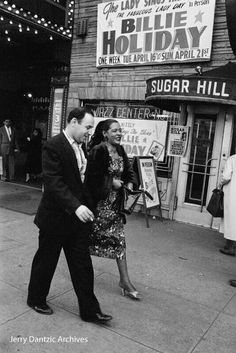 a man and woman walking down the sidewalk in front of a building with signs on it