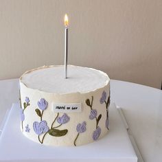 a white cake with purple flowers and a single candle on top is sitting on a table