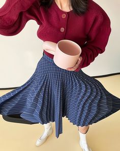 a woman in a red sweater and blue pleated skirt holding a pink coffee cup