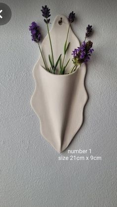 a white vase with purple flowers in it hanging on the wall next to a gray wall