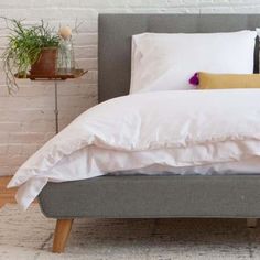 a bed with white sheets and pillows in front of a brick wall next to a potted plant