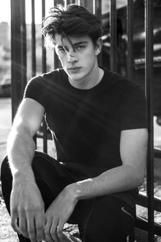 black and white photograph of a young man sitting on the ground in front of a fence