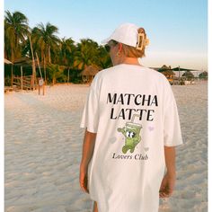 a woman standing on top of a sandy beach next to the ocean wearing a t - shirt that says matcha latte