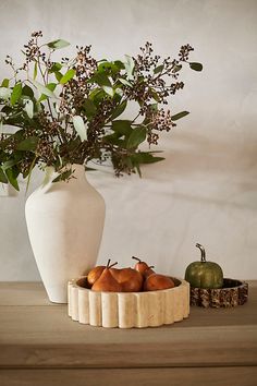 a white vase filled with lots of fruit on top of a wooden table next to a potted plant