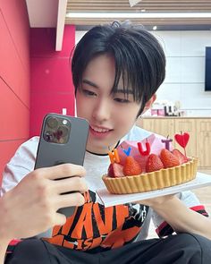 a young man holding a cell phone and looking at a cake on a plate with strawberries